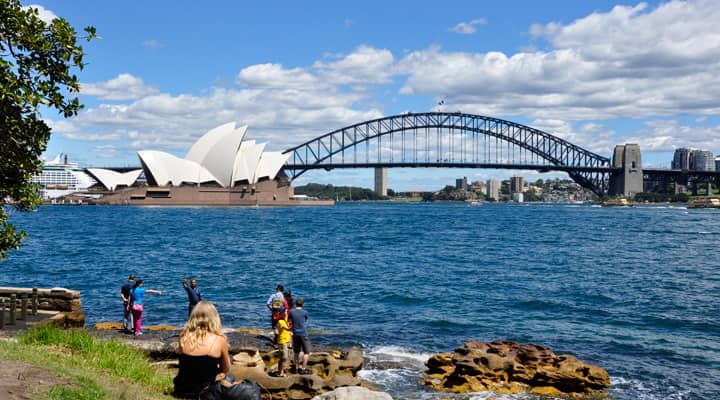 A Sydney City tour bus has stopped at Mrs Macquaries Point, a popular lookout offering stunning views of the Sydney Opera House and Sydney Harbour Bridge. Tourists are enjoying the iconic views and taking pictures.