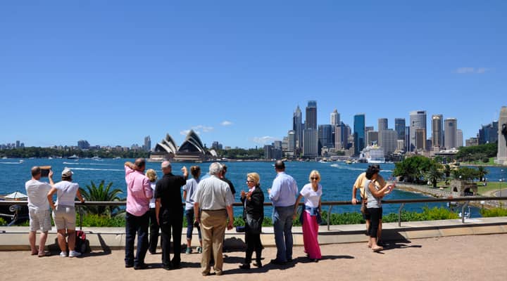 A Sydney sightseeing bus has stopped at the Gap, a popular lookout in Watsons Bay, Australia, offering stunning views of Sydney Harbour and the ocean. Tourists are enjoying the panoramic scenery and taking pictures.