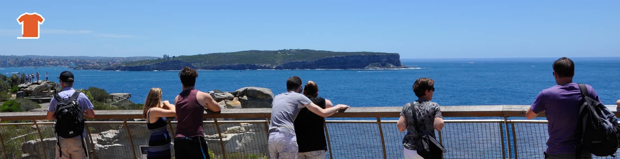 A group of tourists stand at The Gap lookout in Sydney, Australia, captivated by the breathtaking view. The vast expanse of the Tasman Sea stretches before them, marking the entrance to Sydney Harbour. North Head and the North Shore rise majestically in the distance.