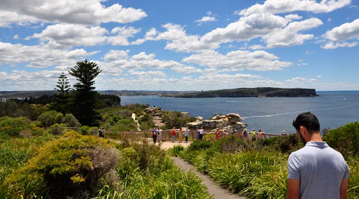 A Sydney sightseeing bus has stopped at the Gap, a popular lookout in Watsons Bay, Australia, offering stunning views of Sydney Harbour and the ocean. Tourists are enjoying the panoramic scenery and taking pictures.