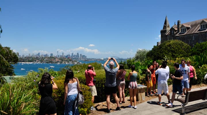 A group of tourists on a Sydney sightseeing bus tour enjoy the views of the city skyline from the lookout next to Kincoppal School in Vaucluse, Sydney, Australia. The tourists are taking pictures of the city, which is in the distance.