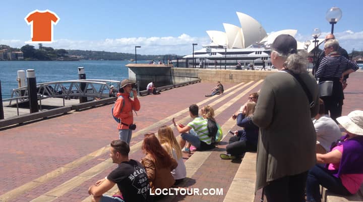 A group of tourists on a Sydney and The Rocks walking tour are gazing out at Sydney Harbour with the iconic Sydney Opera House in the background. A tour guide in a bright orange uniform points out landmarks and shares stories of early Sydney.