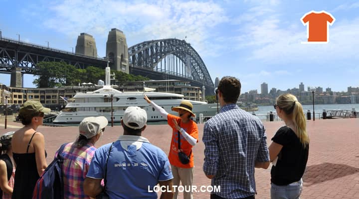 A tour guide, dressed in a bright orange uniform, stands at Campbells Cove explaining the history of Sydney's historic Rocks precinct to a group of tourists. The iconic Sydney Harbour Bridge arches over the picturesque scene.