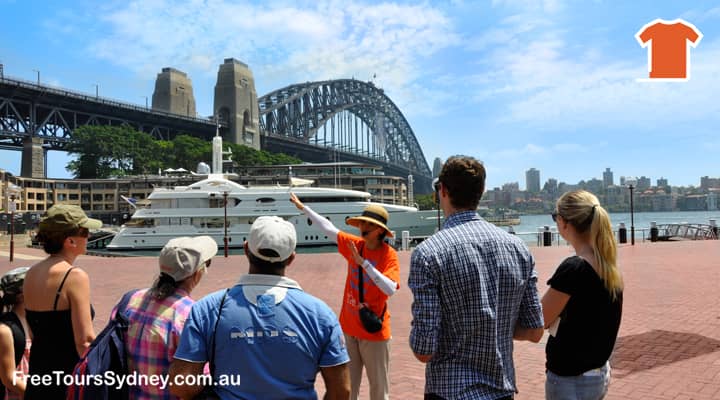 A local tour guide in an orange uniform is giving a walking tour of the Rocks, the oldest surviving settlement in Sydney. The Sydney Harbour Bridge is in the background of the picture. The tour guide is pointing out some of the historical landmarks in the area, such as the Cadman's Cottage and the Woolstores, and telling stories about the history of the Rocks. The tourists are listening attentively.