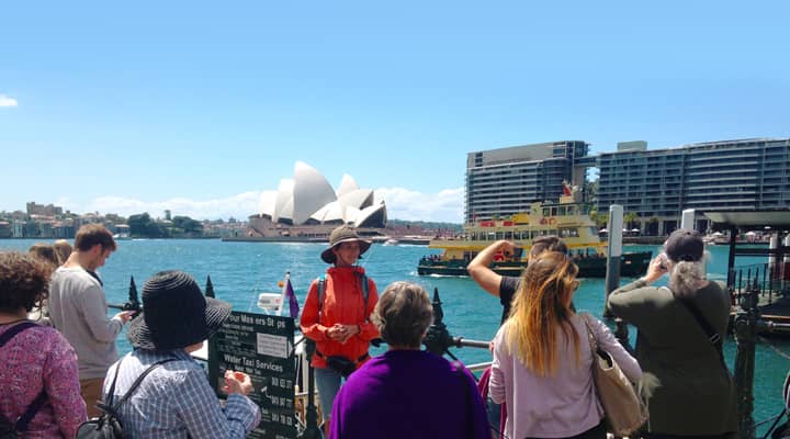 A group of tourists on a Free Sydney walking tour are gazing the iconic Sydney Opera House in the background. A tour guide in a bright orange 'free tours' uniform.