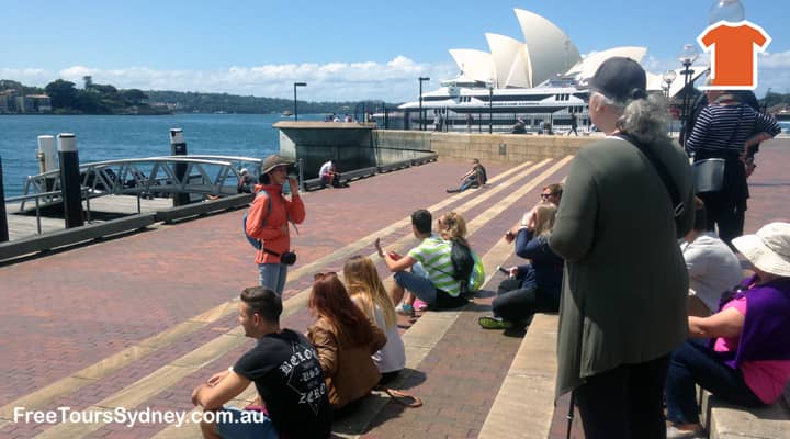 A group of tourists are enjoying a Sydney walking tour. They are standing on the waterfront, looking out at Sydney Harbour. The Sydney Opera House is in the background. A tour guide in an orange 'free tours' uniform is pointing out some of the landmarks and telling stories of early Sydney.