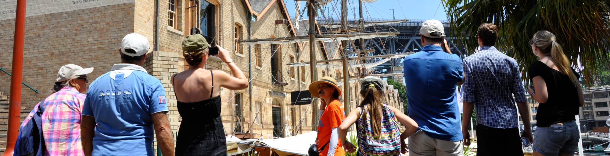 A tour guide in free tours Sydney uniform is leading a group of tourists in The Rocks, Sydney. Old woolstores and sails of a ship in the background.