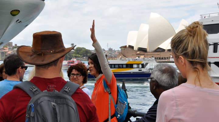 A group of tourists on a Free Sydney walking tour are gazing the iconic Sydney Opera House in the background. A tour guide in a bright orange 'free tours' uniform.